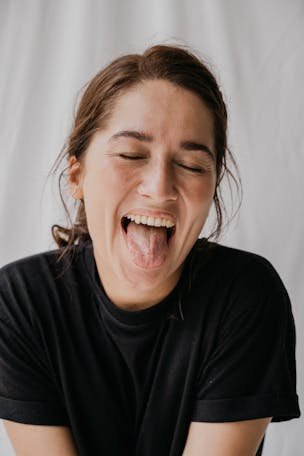 A joyful woman in a black shirt playfully sticks out her tongue with eyes closed against a soft white background.