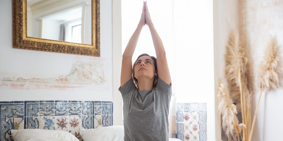 A woman begins her day with yoga, sitting on a bed in a serene, well-decorated bedroom.