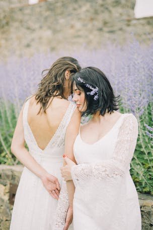 Two women in wedding dresses share a tender moment in a blooming lavender field.