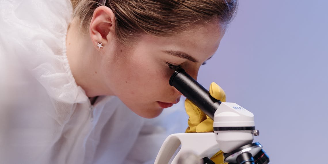 A focused female scientist studies samples under a microscope in a laboratory setting, wearing protective gear.