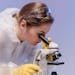 A focused female scientist studies samples under a microscope in a laboratory setting, wearing protective gear.