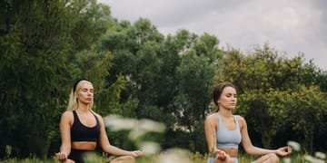 Two women practicing yoga meditation in a serene outdoor park setting.